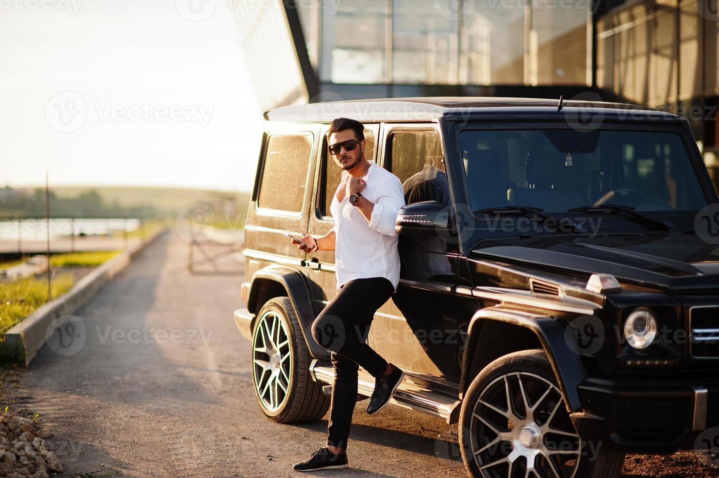 un hombre asiático sólido con camisa blanca y gafas de sol posó cerca de un camión suv de la mafia negra. foto