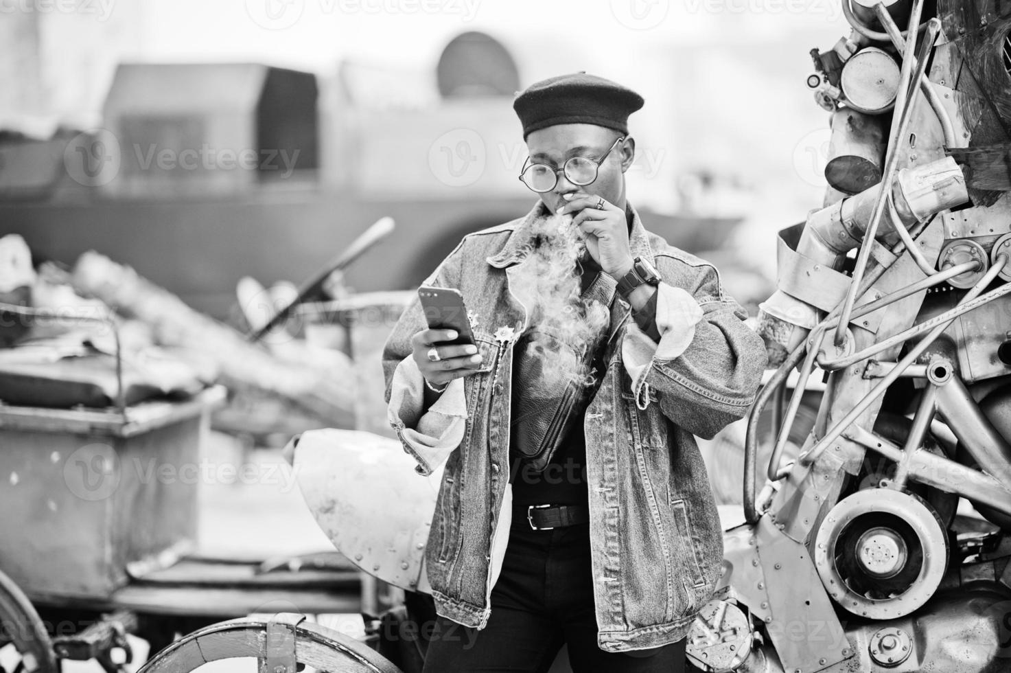 African american man in jeans jacket, beret and eyeglasses, smoking cigar and posed against old retro vehicle and looking at phone. photo
