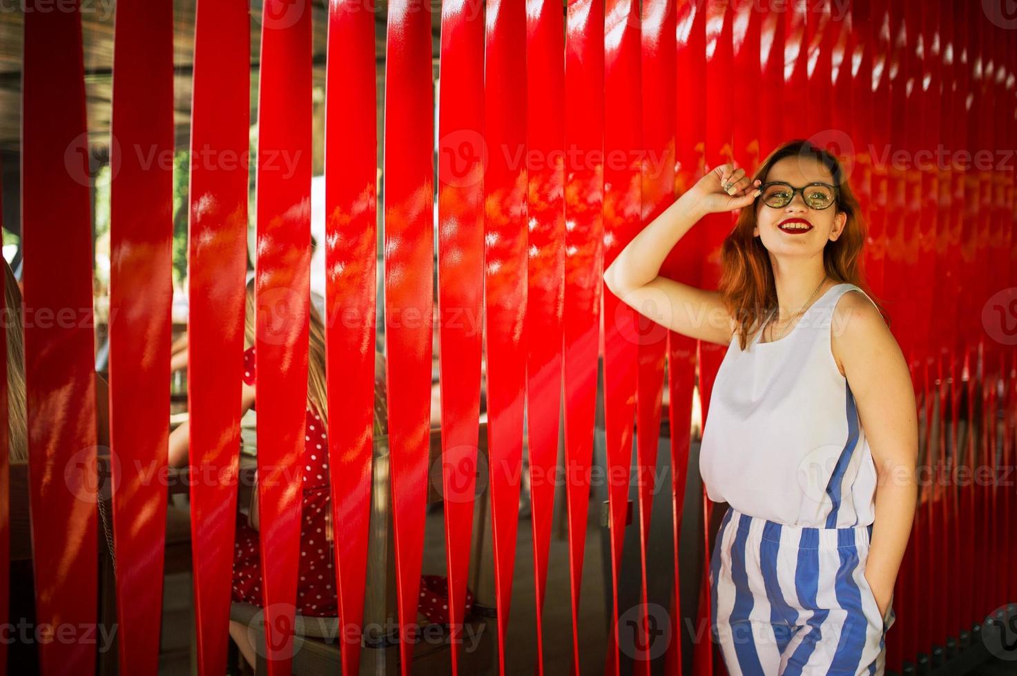 Attractive redhaired woman in eyeglasses posing against red background. photo