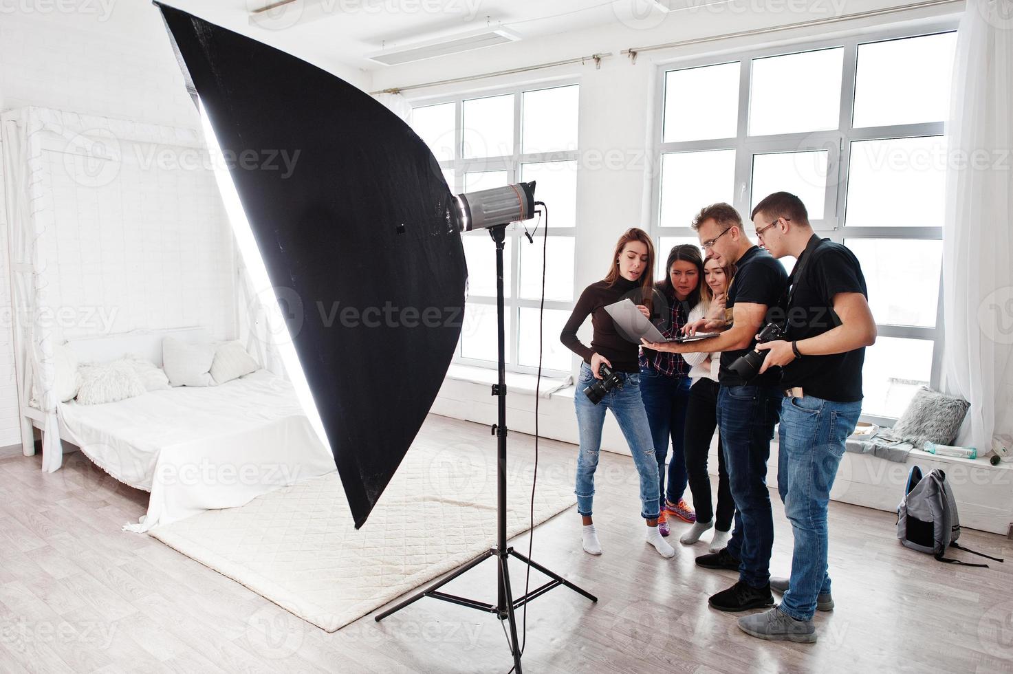 Photographer explaining about the shot to his team in the studio and looking on laptop. Talking to his assistants holding a camera during a photo shoot. Teamwork and brainstorm.