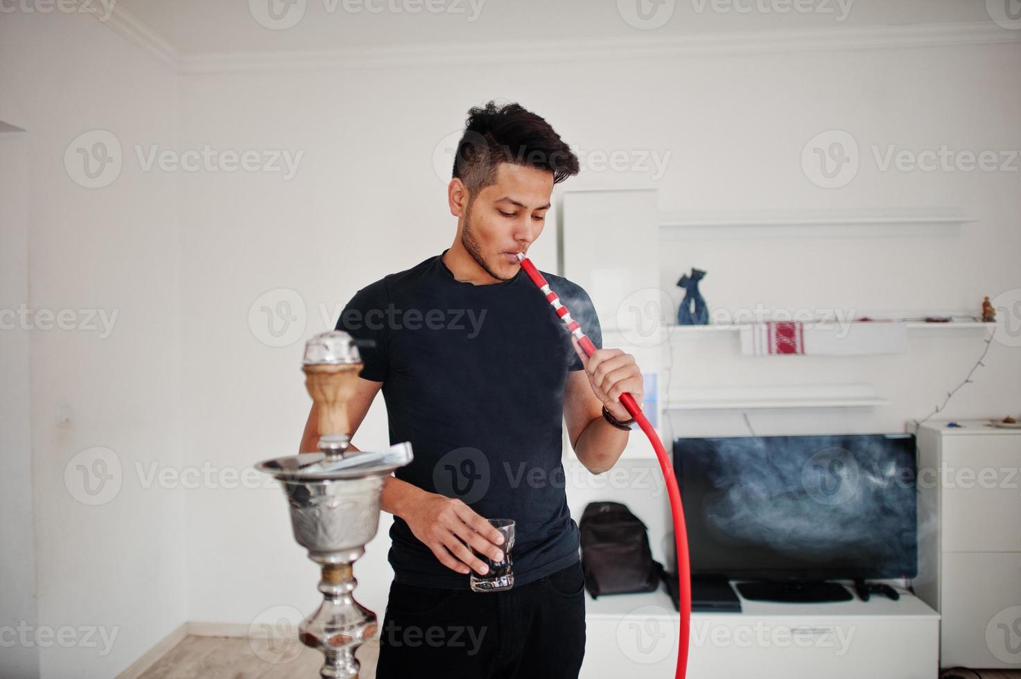 Handsome and fashionable indian man in black standing at room, smoking hookah with glasses at hand. photo