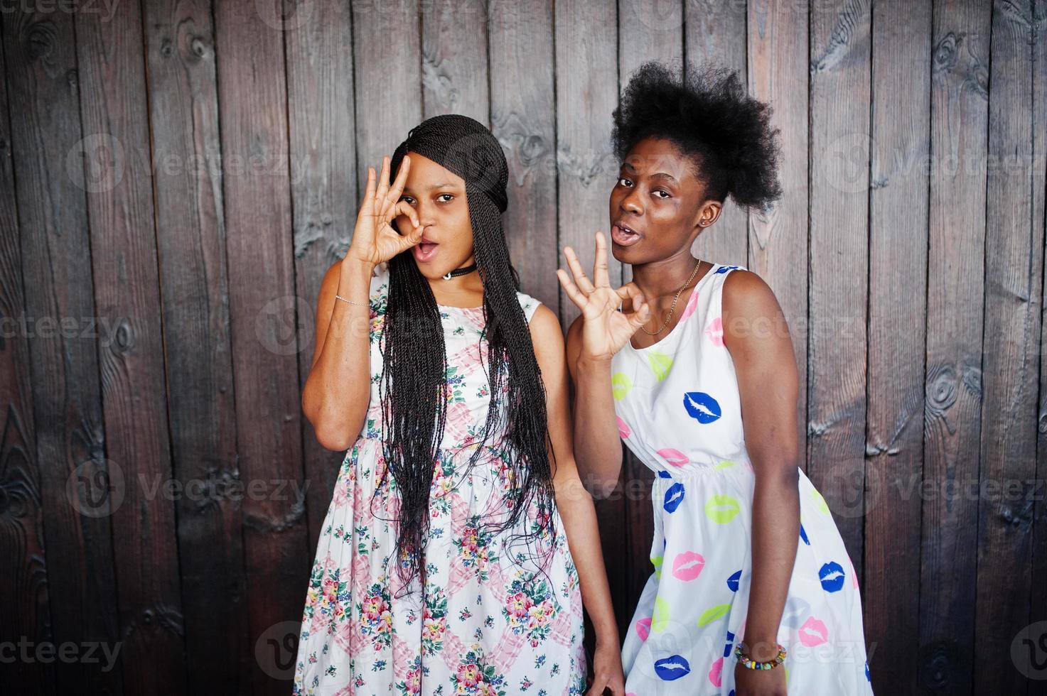 Two black african girlfriends at summer dresses posed against dark wooden background and showing okay fingers sign. photo