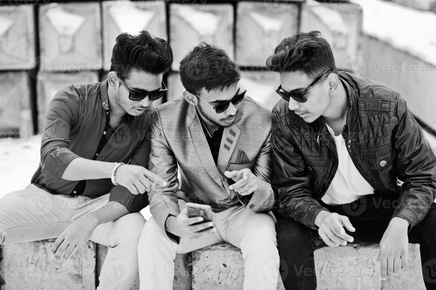 Group of three casual young indian mans in sunglasses posed against stone blocks and looking at mobile phone. photo