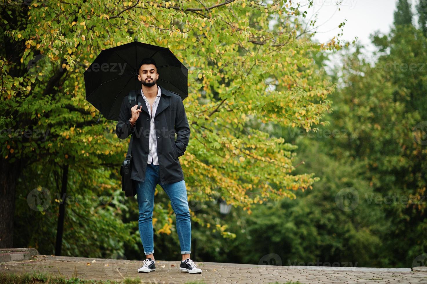 Fashionable tall arab beard man wear on black coat with umbrella and bag case posed at rain weather day. photo