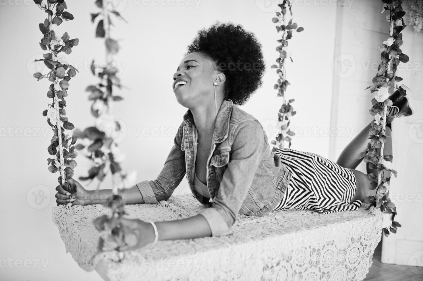 Attractive african american woman with afro hair wear on skirt and jeans jacket, posed at white room on swing. Fashionable black model. photo