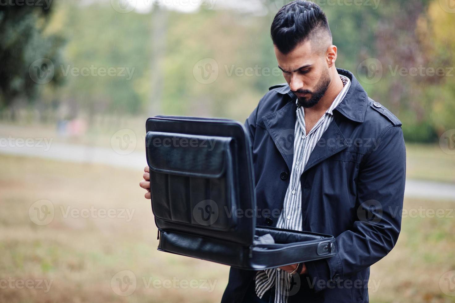 Fashionable tall arab beard man wear on black coat with bag case posed at rain weather day. photo