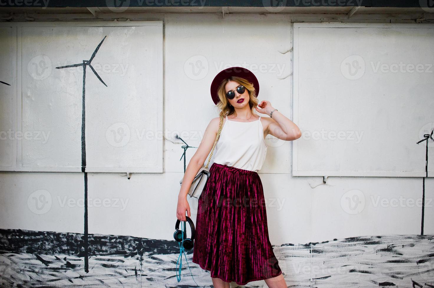 Chica modelo rubia de moda y hermosa con elegante falda de terciopelo rojo, blusa blanca, sombrero y gafas de sol, posada con teléfono y auriculares. foto