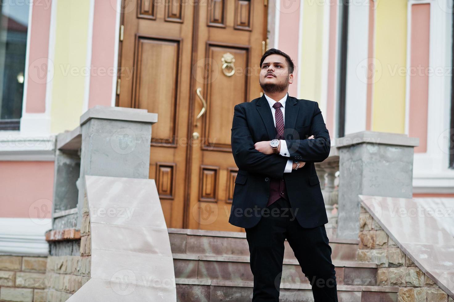 Stylish indian businessman in formal wear standing against door in business center with cross arms. photo