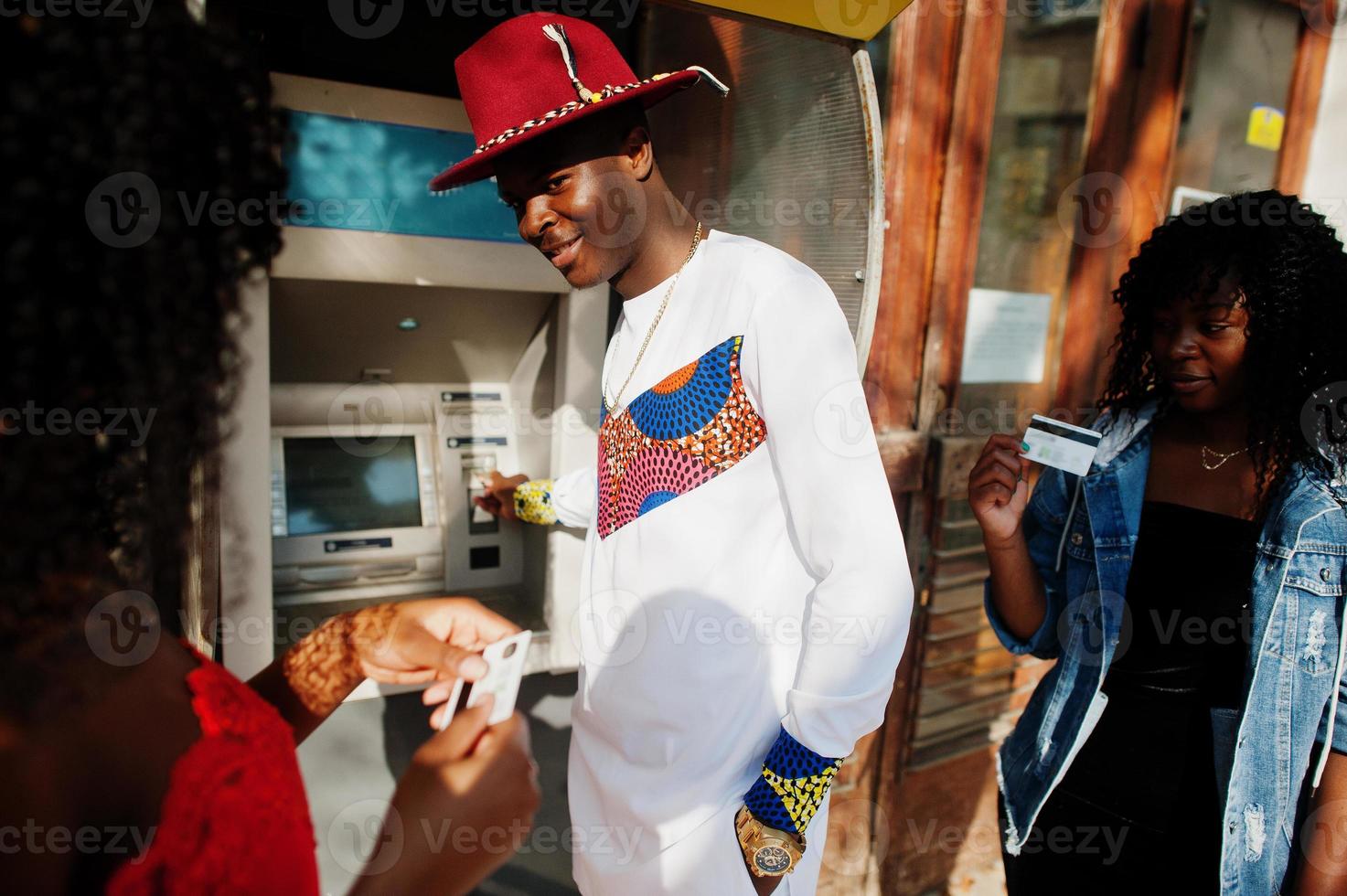 Group of three stylish trendy afro france friends posed against atm. Black african man model with two dark skinned womans with credit cards. photo