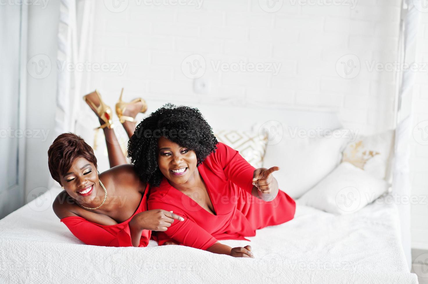 Two fashion african american models in red beauty dress and golden high heels, sexy womans posing evening gown lie on the bed. photo