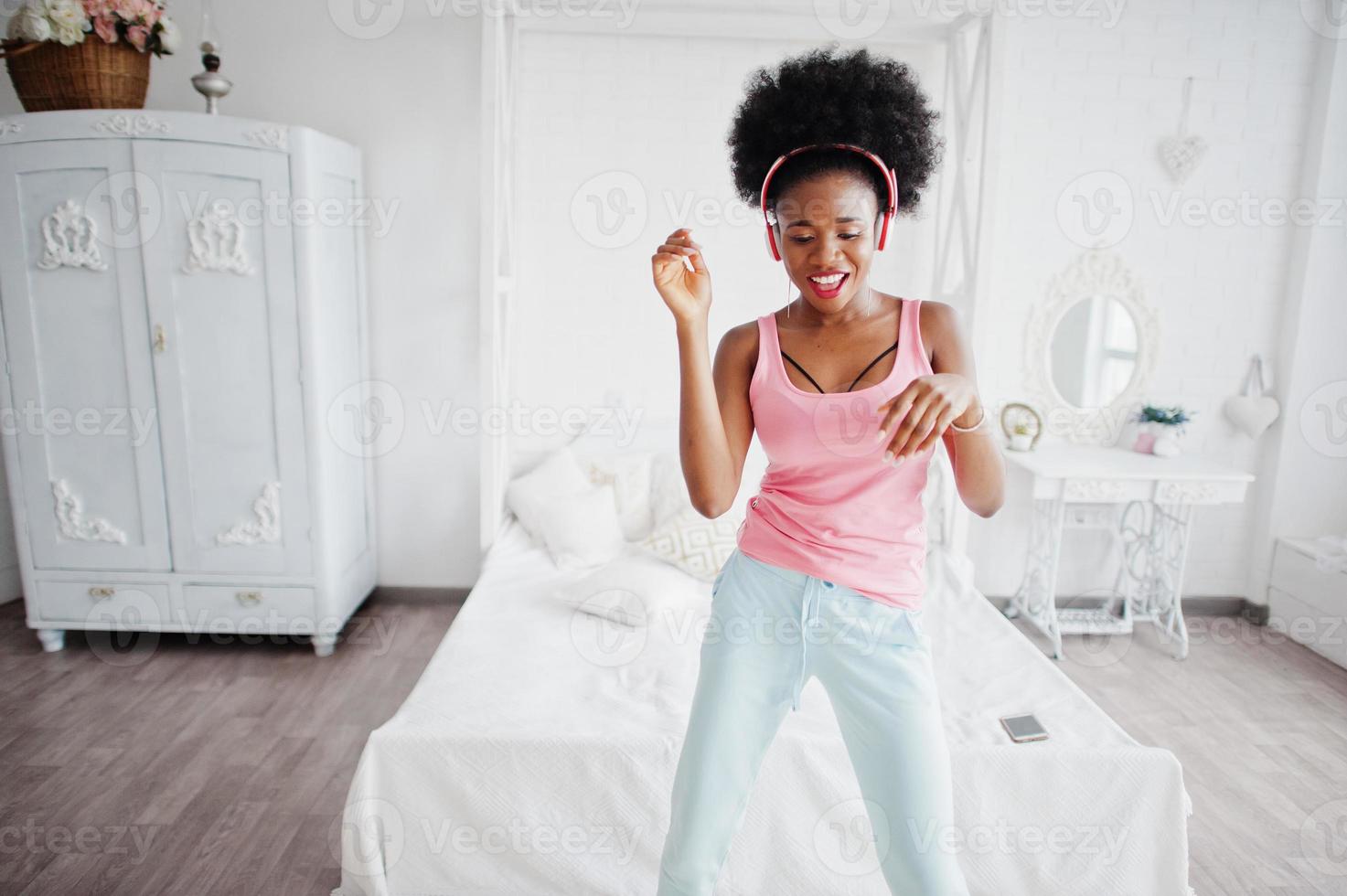 Young african american woman in pink singlet dancing and listen music on earphones at her white room. photo