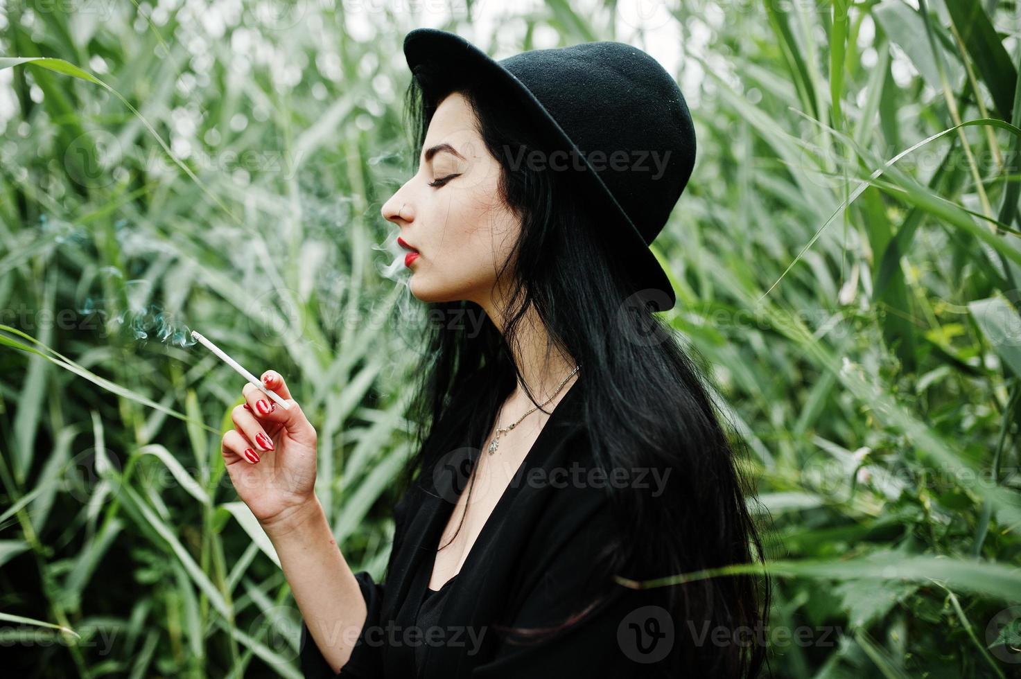 Sensual smoker girl all in black, red lips and hat. Goth dramatic woman smoking on common reed. photo