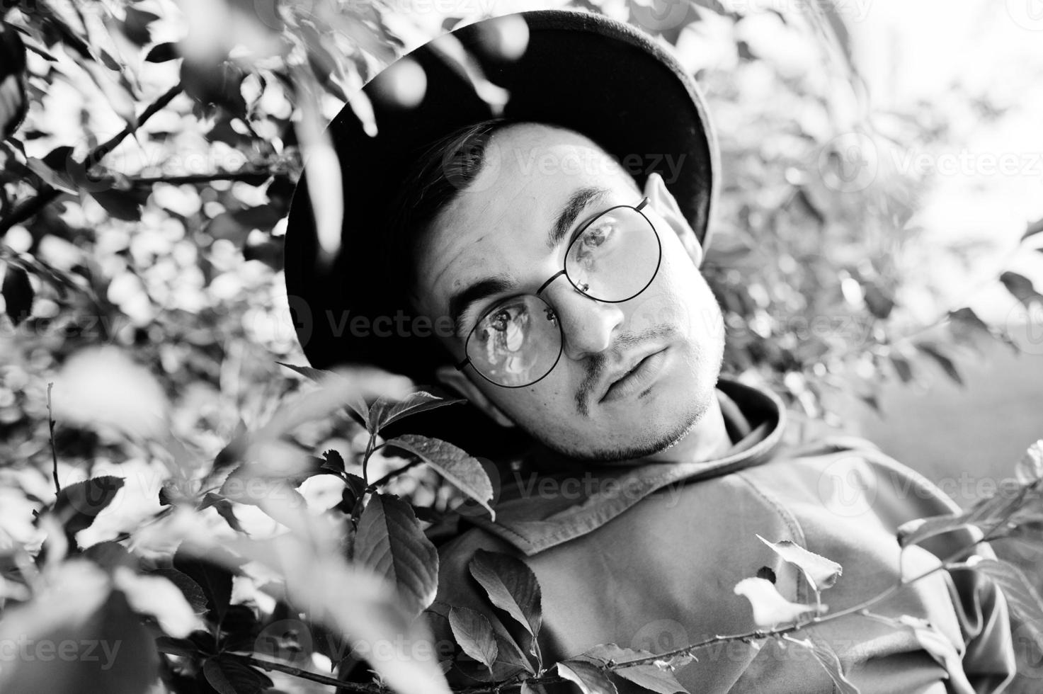 Stylish man in glasses, brown jacket and hat posed on green field. photo