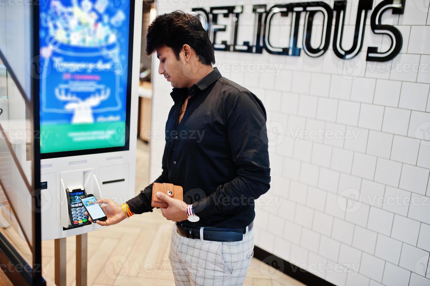 Indian man customer at store place orders and pay by contactless credit card on mobile phone through self pay floor kiosk for fast food, payment terminal. Pay pass. photo