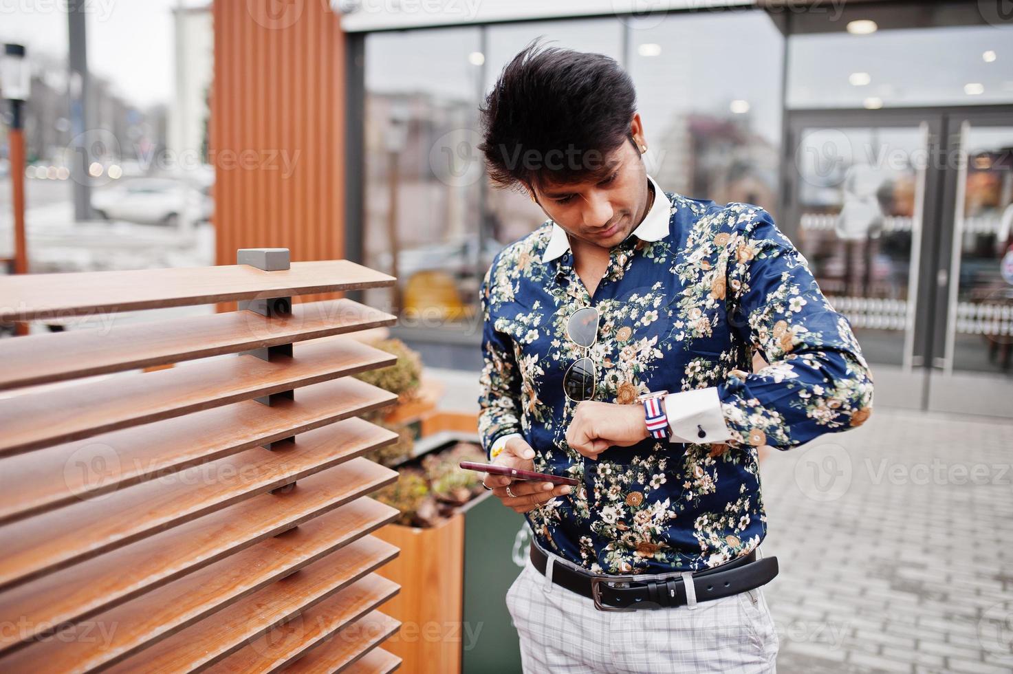 Attractive and handsome stylish indian man wear on shirt, holding mobile phone, looking at watches and standing in the terrace of modern lounge cafe at his work break. photo