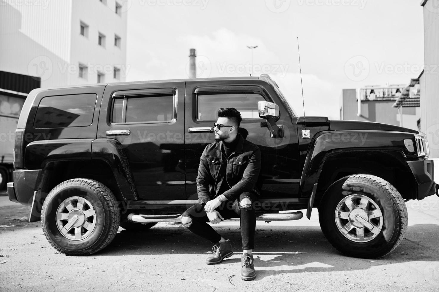 Fashion rich beard Arab man wear on black jeans jacket and sunglasses posed against big black suv car. Stylish, succesful and fashionable arabian model guy. photo