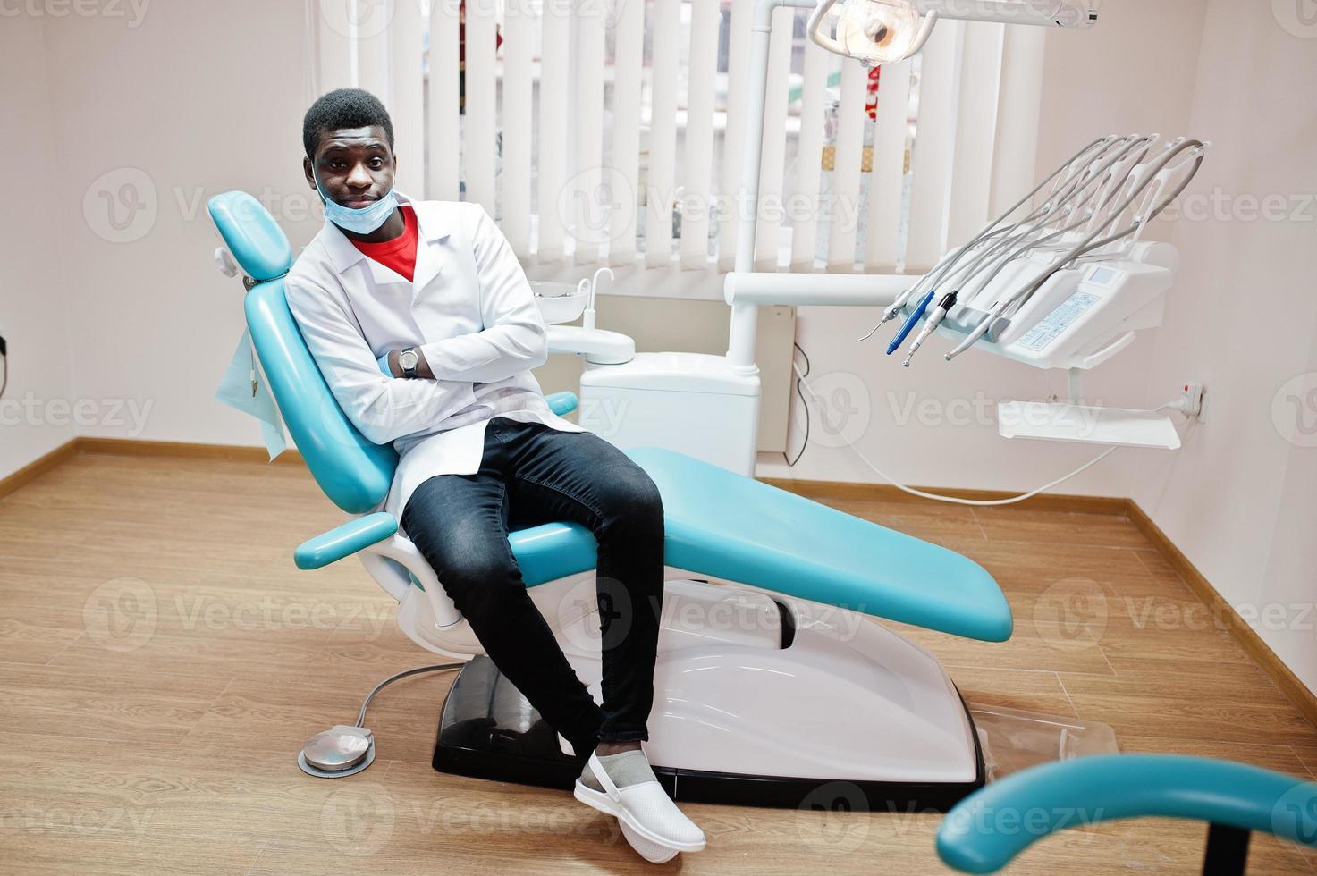 médico afroamericano enmascarado con brazos cruzados sentado en la silla del dentista en la clínica dental. foto