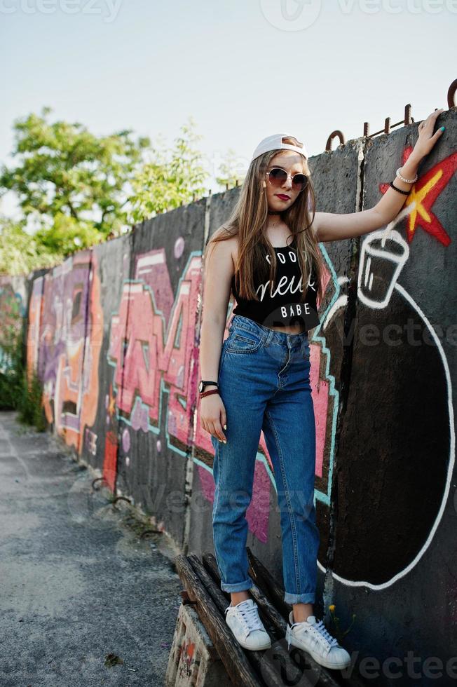 elegante chica hipster casual con gorra, gafas de sol y jeans desgastados contra una gran pared de graffiti con una gran bomba tnt. foto