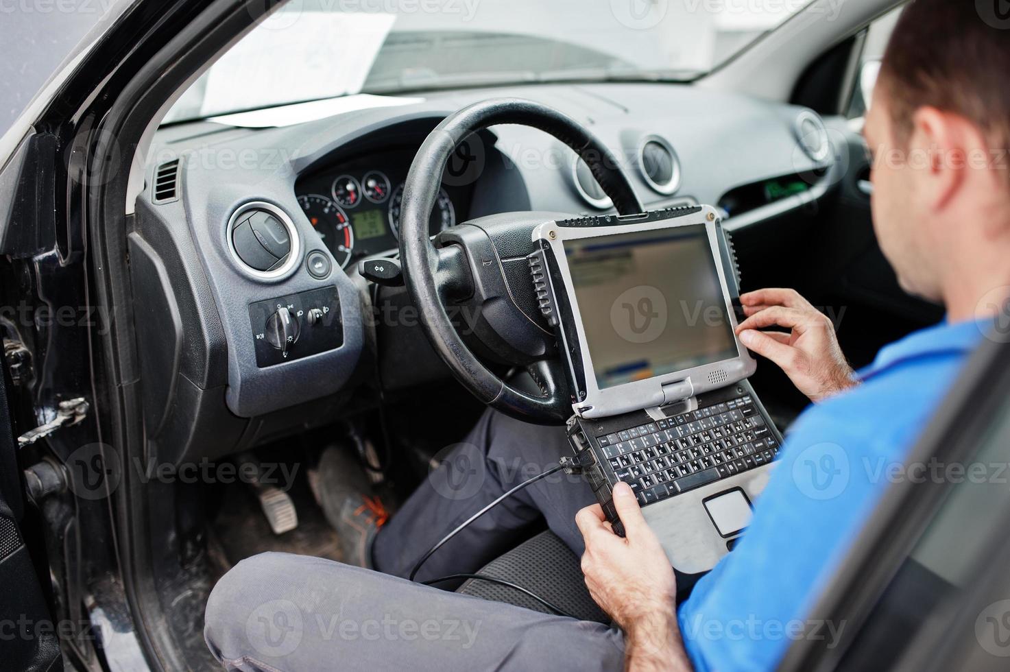 Car repair and maintenance theme. Electric mechanic in uniform working in auto service, making car diagnostics using obd device with laptop. photo