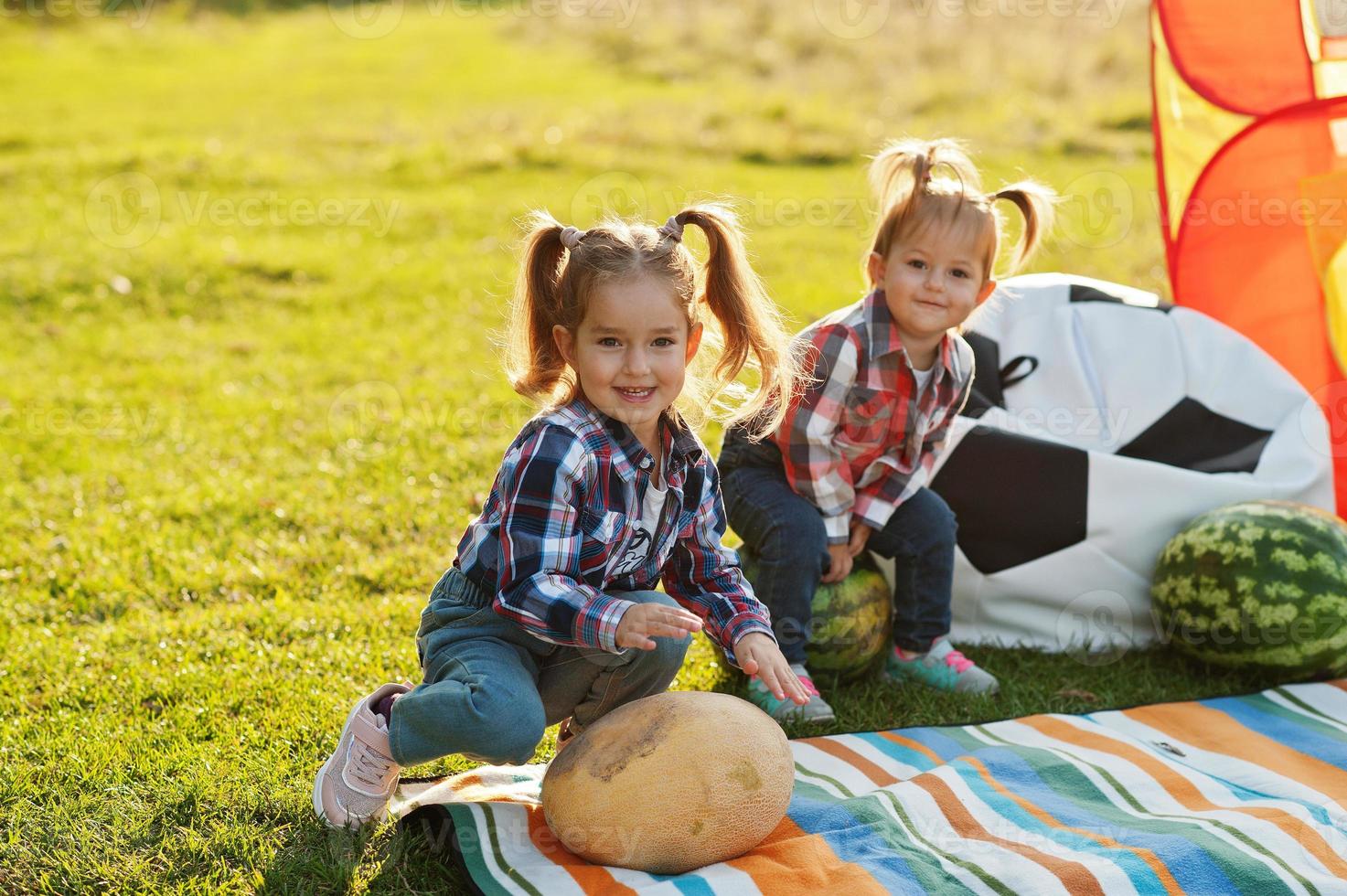 Two little cutie sister girls outdoor in picnic, wear checkered shirt. Sitting on watermelon. photo