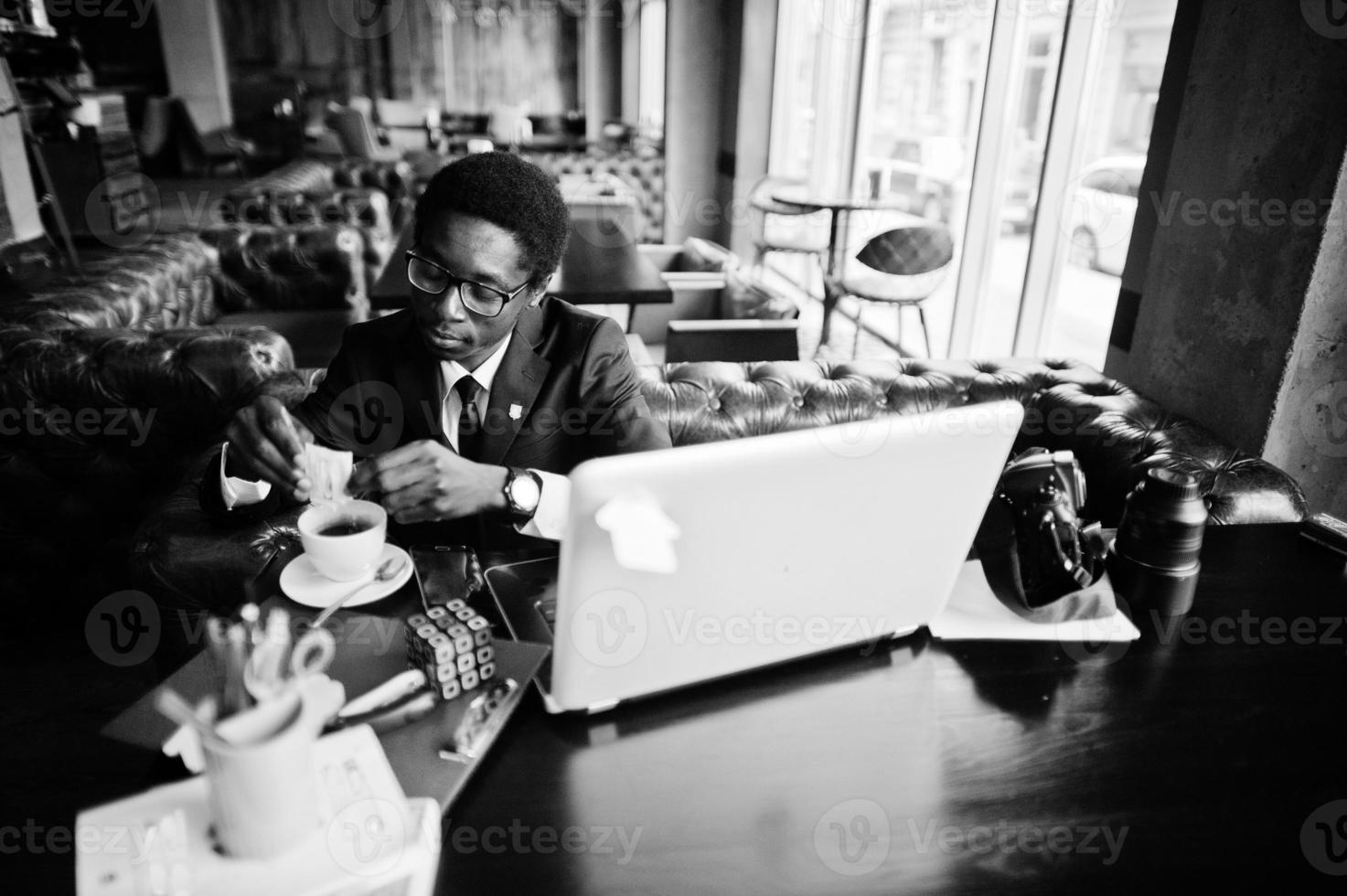 Business african american man wear on black suit and glasses sitting at office with laptop and working, pours sugar into coffee. photo