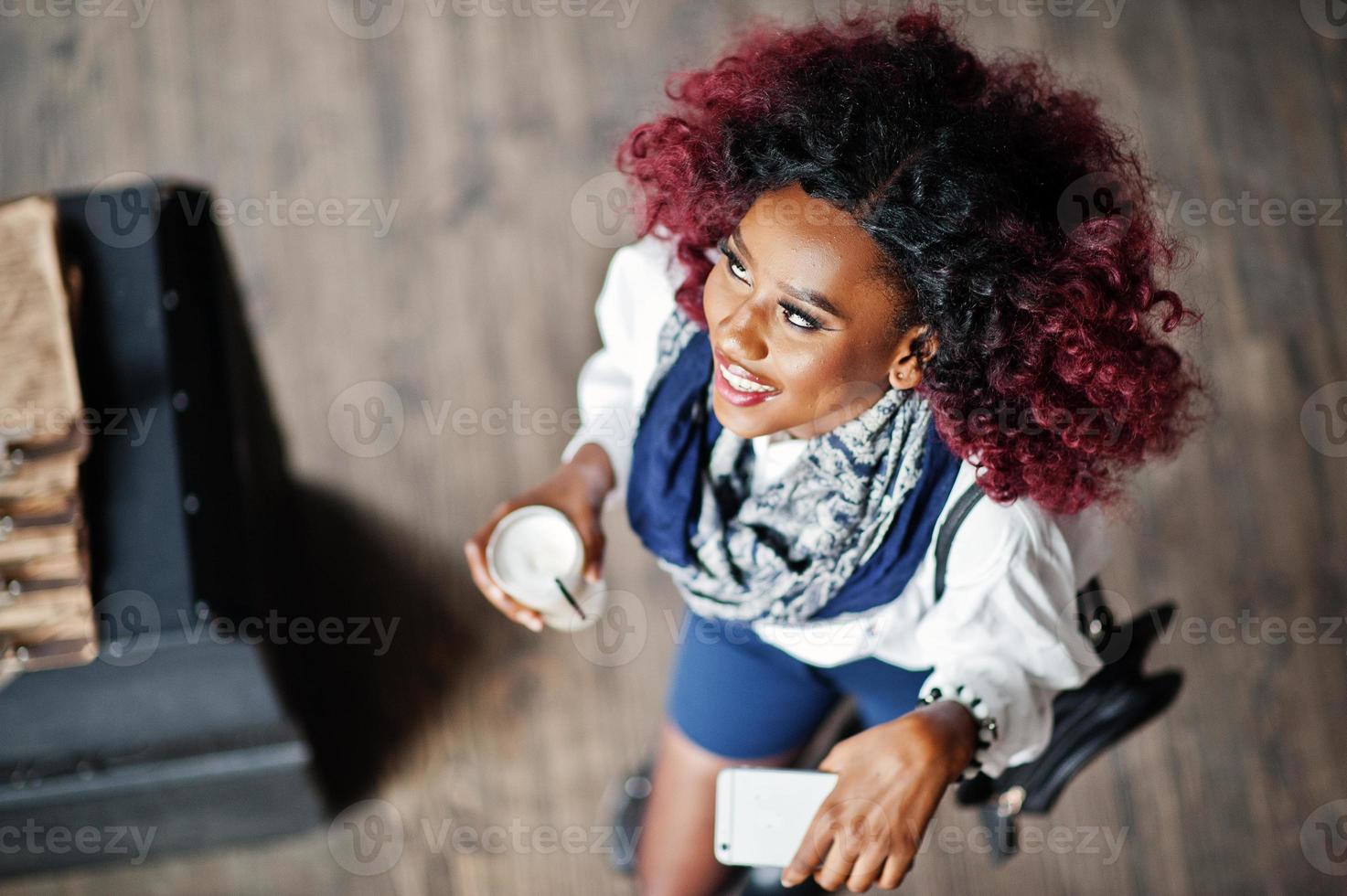 Attractive african american curly girl in white blouse and blue shorts posed at cafe with latte and mobile phone at hand. View from above. photo