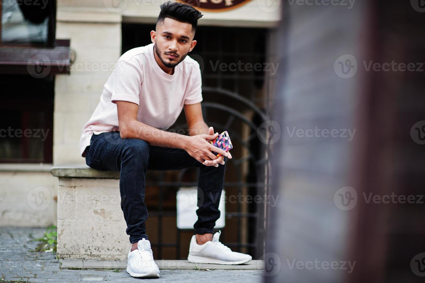 Stylish indian beard man at pink t-shirt. India model sitting with mobile phone at hands outdoor at streets of city. photo