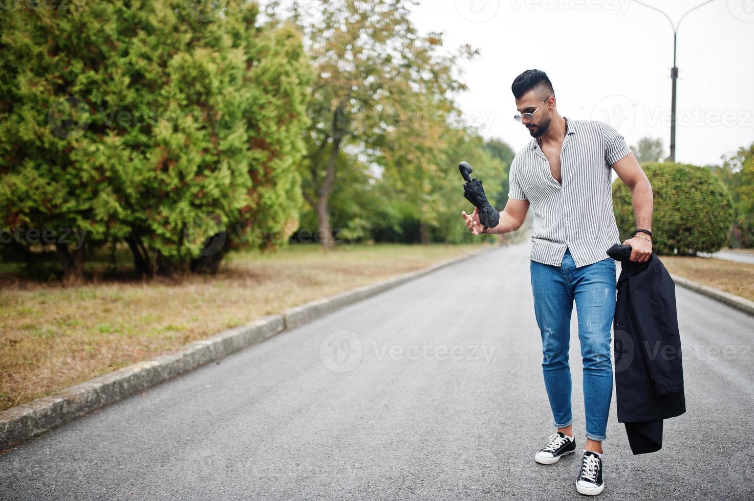 el hombre de barba árabe alto de moda usa camisa, jeans y gafas de sol caminando en el parque con paraguas y abrigo a mano. foto