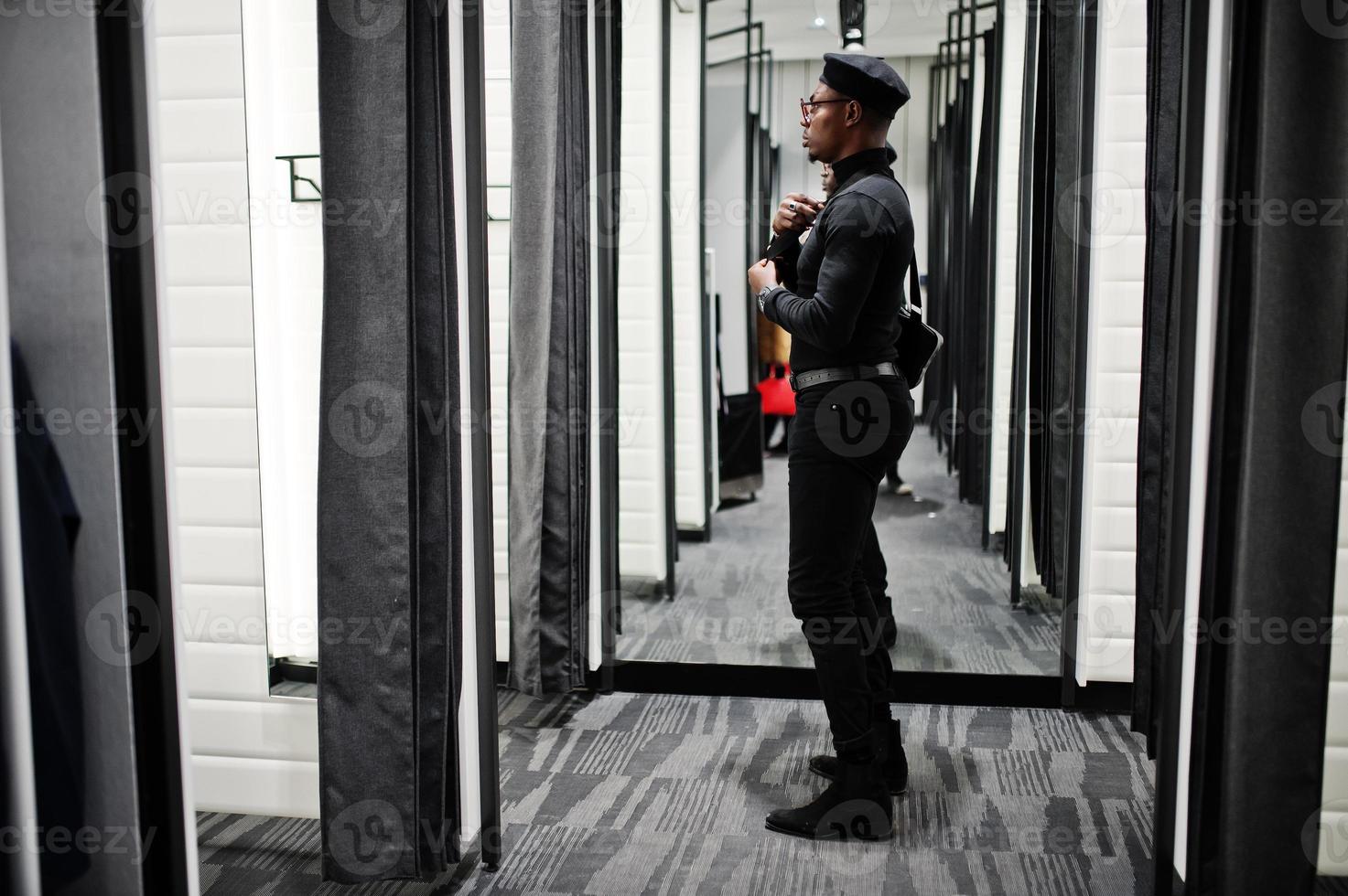 Stylish casual african american man at black outfit and beret with waist bag at fitting room clothes store , looking at mirror. photo