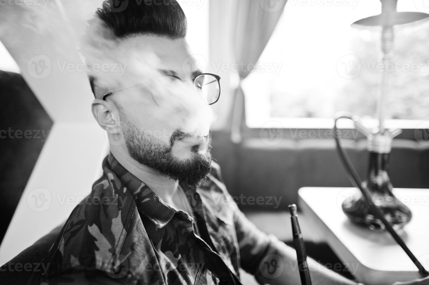 Close up head of stylish beard arabian man in glasses and military jacket with smoke smoking hookah at street bar. Arab model having rest. photo