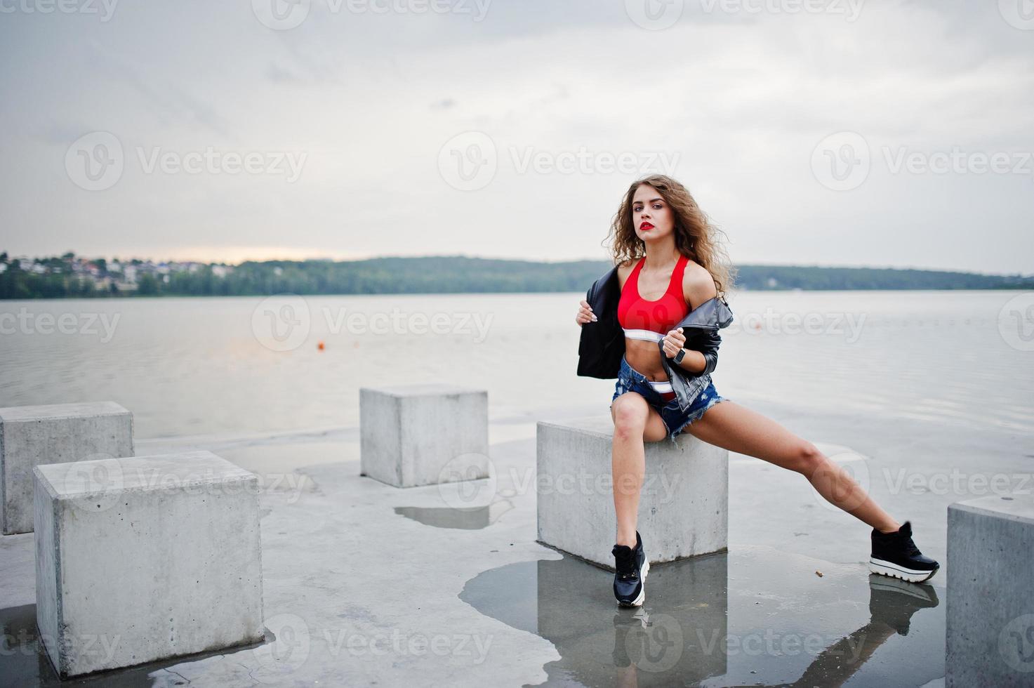 Sexy curly model girl in red top, jeans denim shorts, leather jacket and sneakers posed at stone cubes. photo