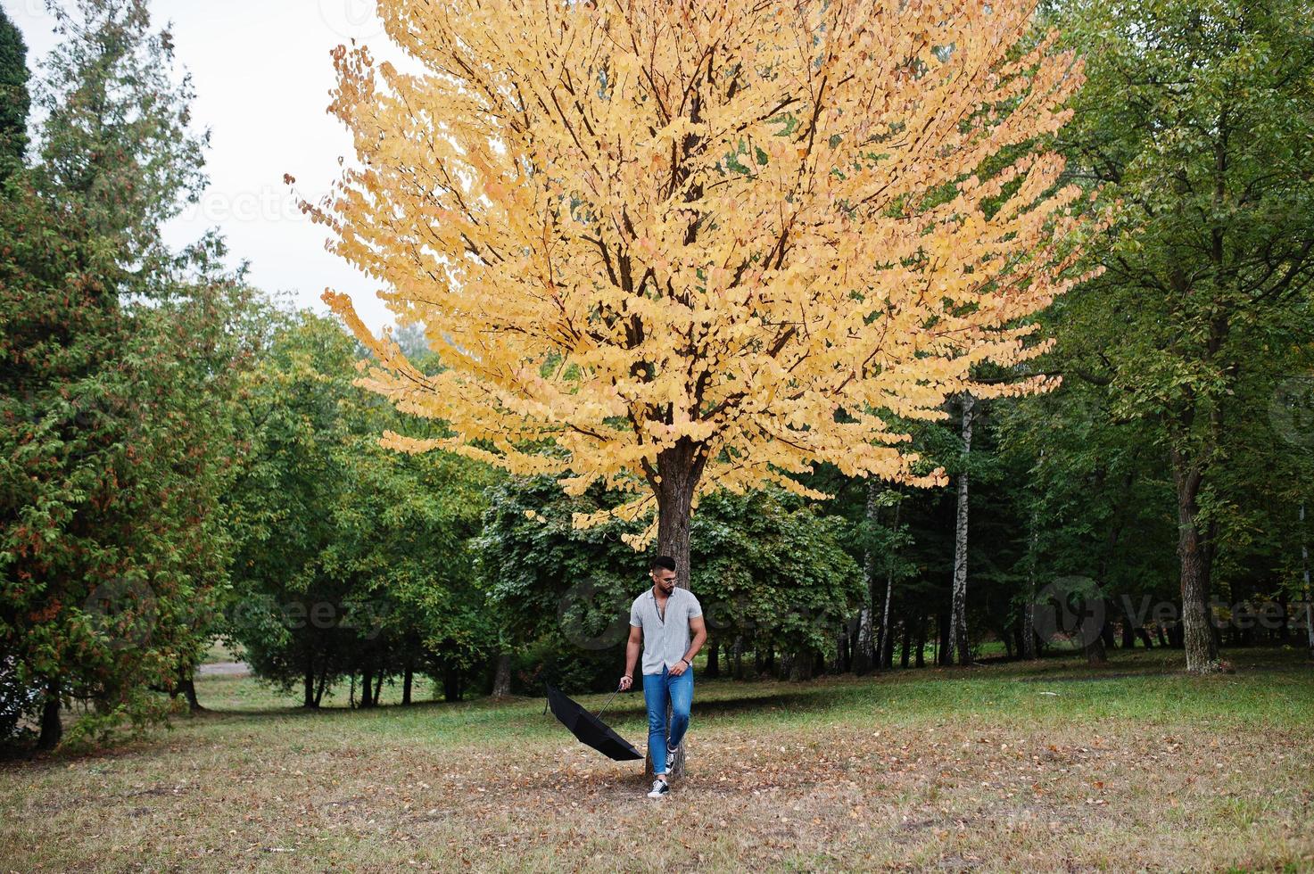 Fashionable tall arab beard man wear on shirt, jeans and sunglasses posed on autumn park with umbrella against yellow leaves tree. photo