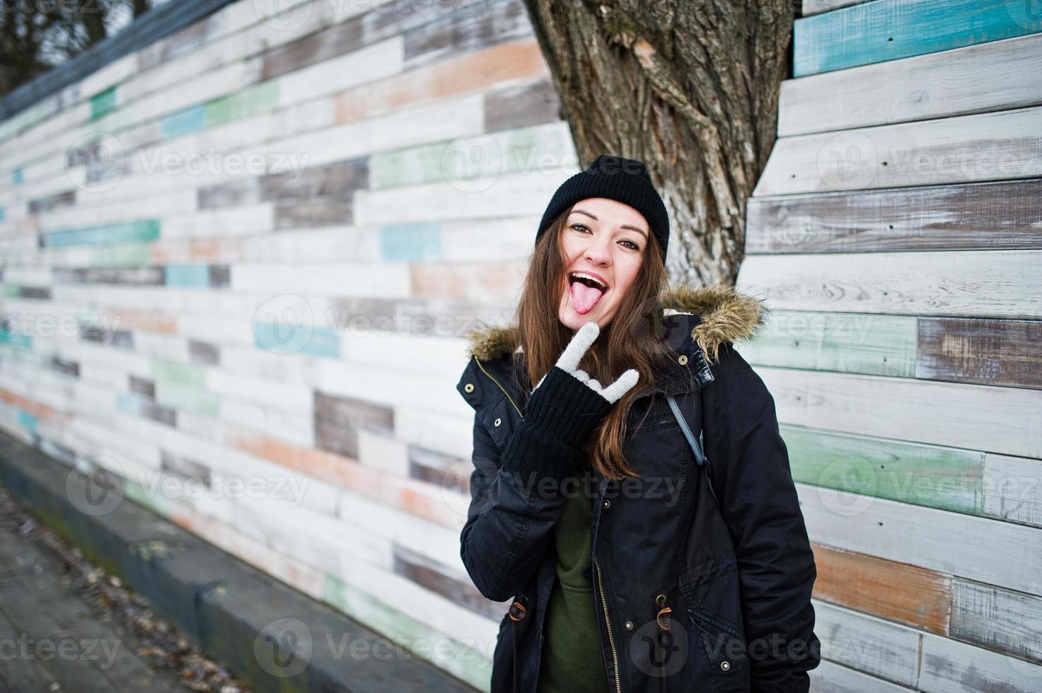 Young girl wear black headwear against wooden wall shows tongue. photo