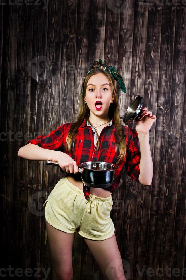 joven ama de casa divertida en camisa a cuadros y pantalones cortos amarillos pin up estilo con cacerola sobre fondo de madera. foto