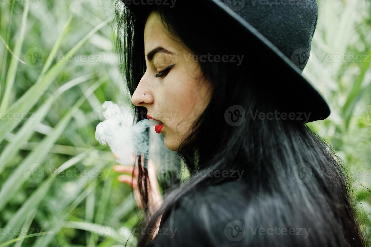 Sensual smoker girl all in black, red lips and hat. Goth dramatic woman smoking on common reed. photo