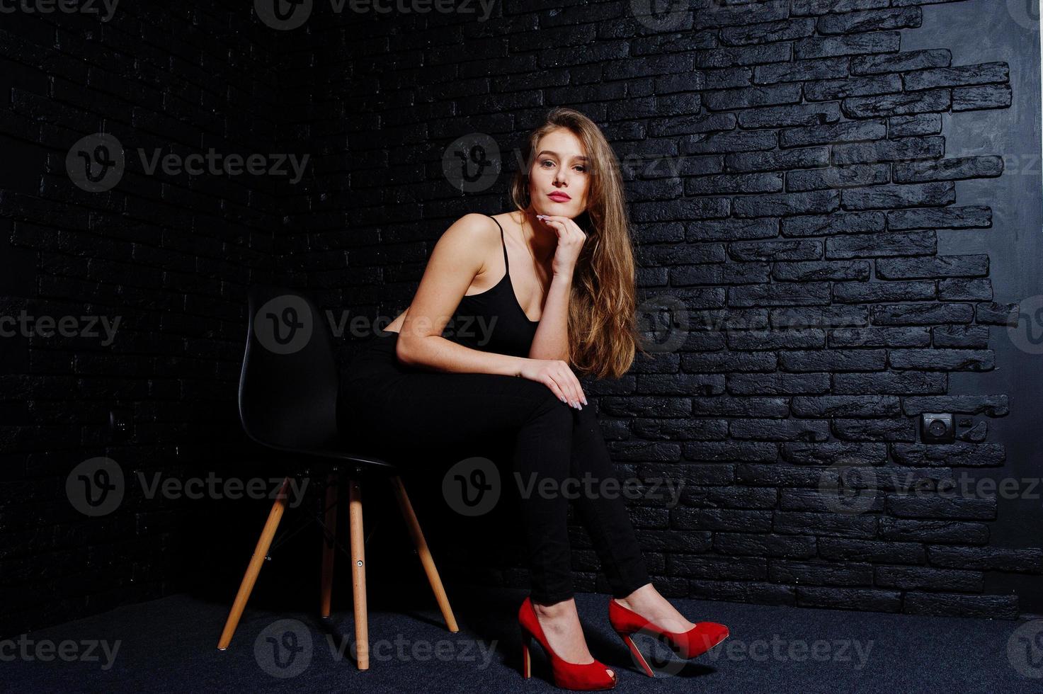 Handsome brunette girl wear on black and red high heels, sitting and posing on chair at studio against dark brick wall. Studio model portrait. photo