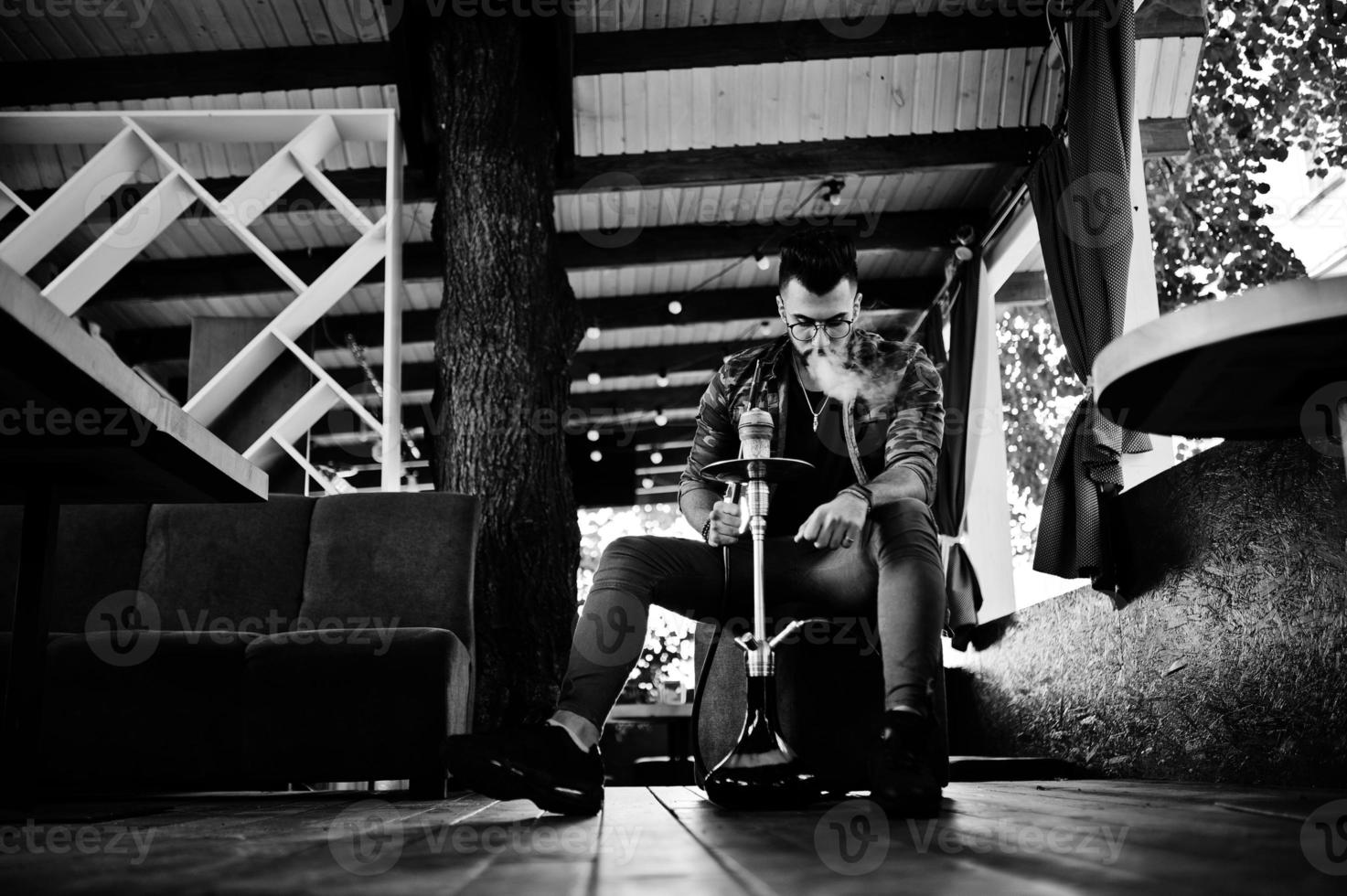 Stylish beard arabian man in glasses and military jacket smoking hookah at street bar. Arab model having rest. photo