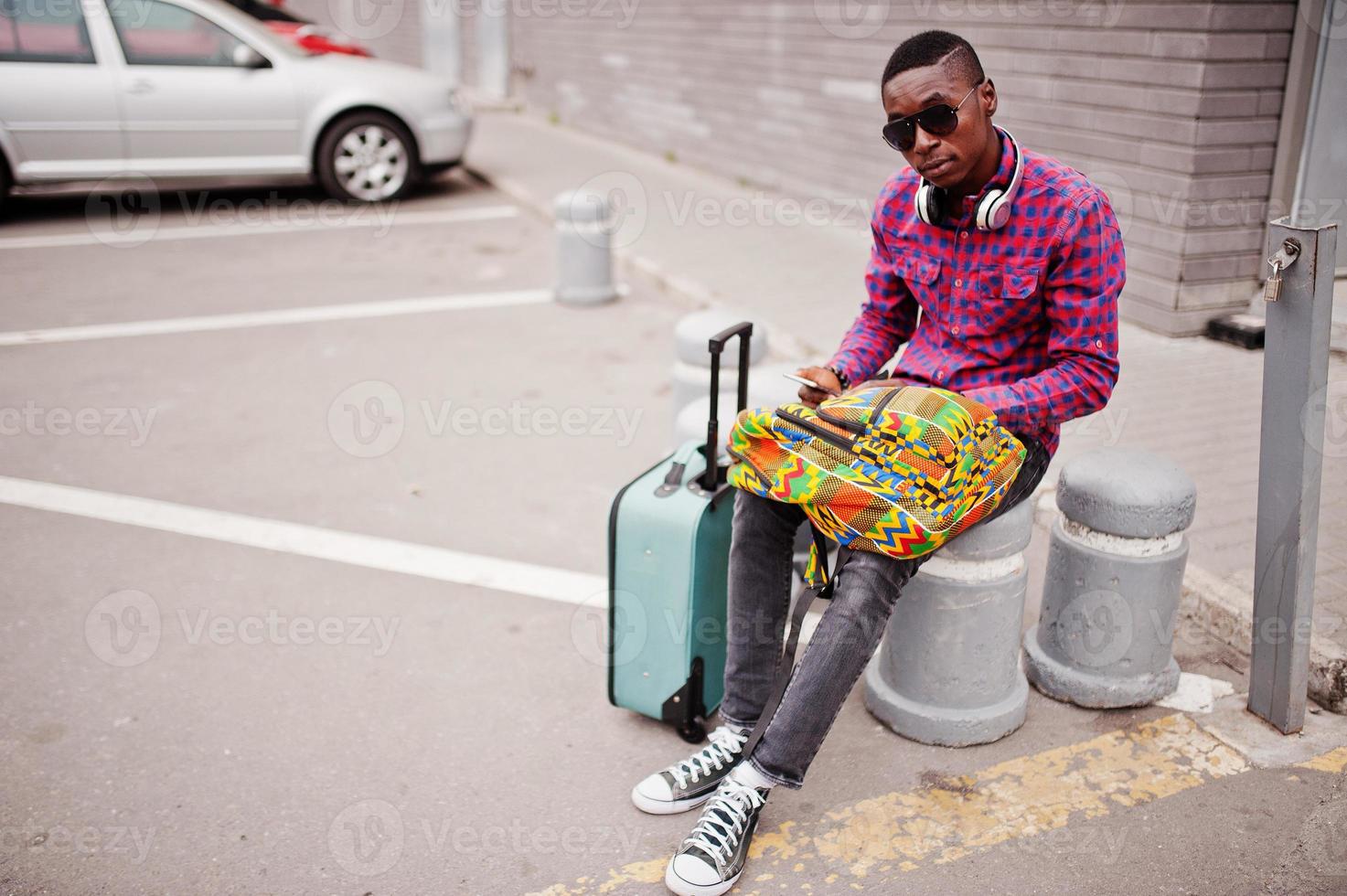 African american man in checkered shirt, sunglasses and earphones with suitcase and backpack. Black man traveler holding mobile phone at hand. photo