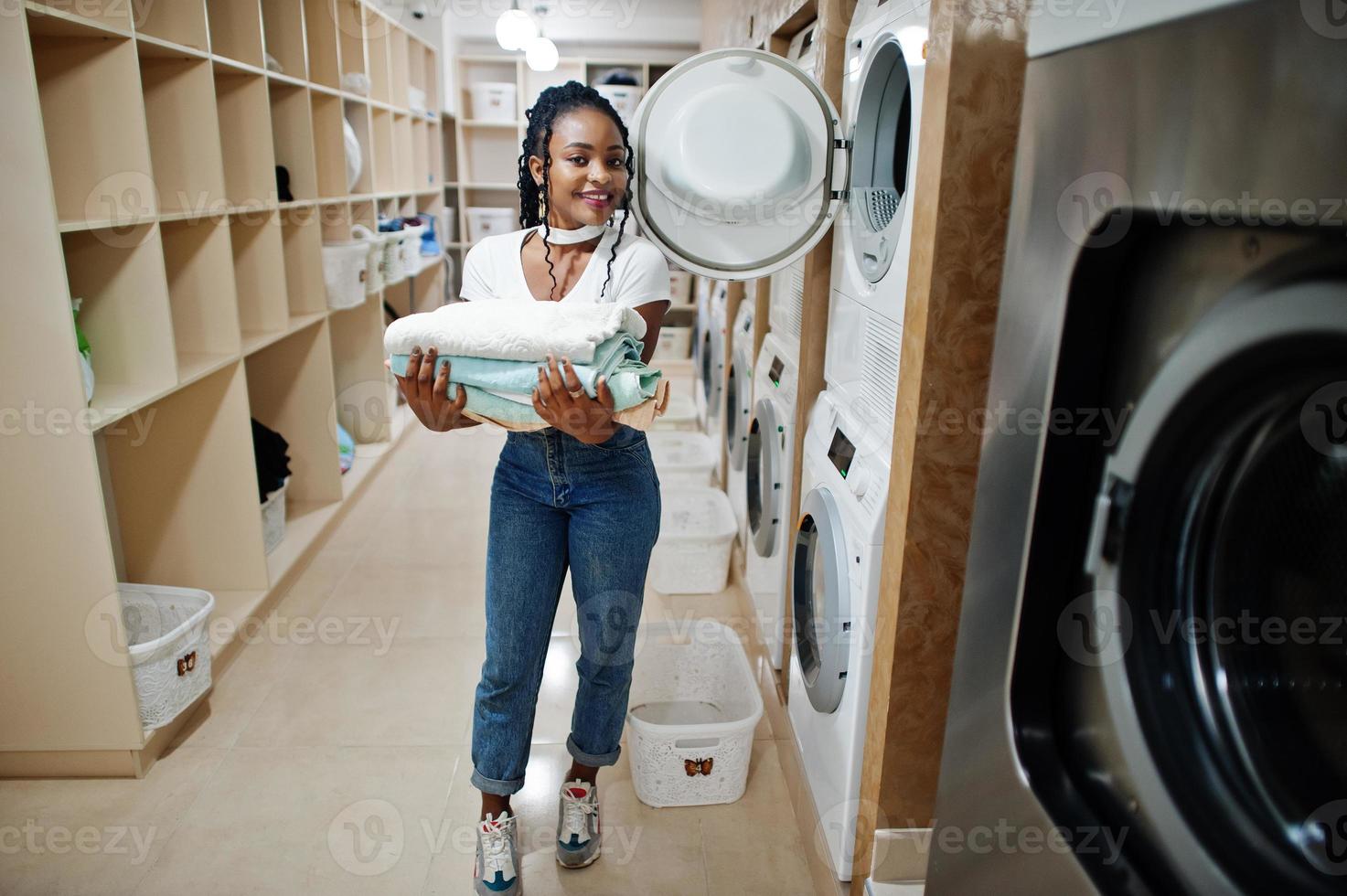 alegre mujer afroamericana con toallas en las manos cerca de la lavadora en la lavandería de autoservicio. foto