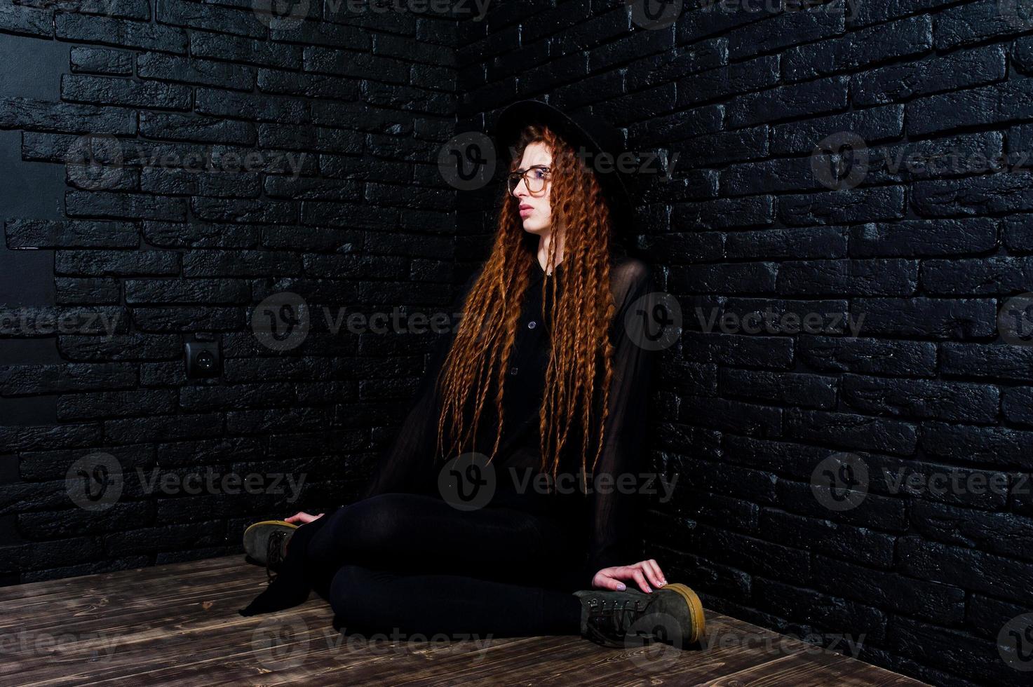 Studio shoot of girl in black with dreads, at glasses and hat on brick background. photo