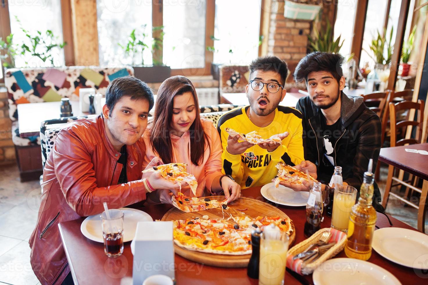 Group of asian friends eating pizza during party at pizzeria. Happy indian people having fun together, eating italian food and sitting on couch. photo