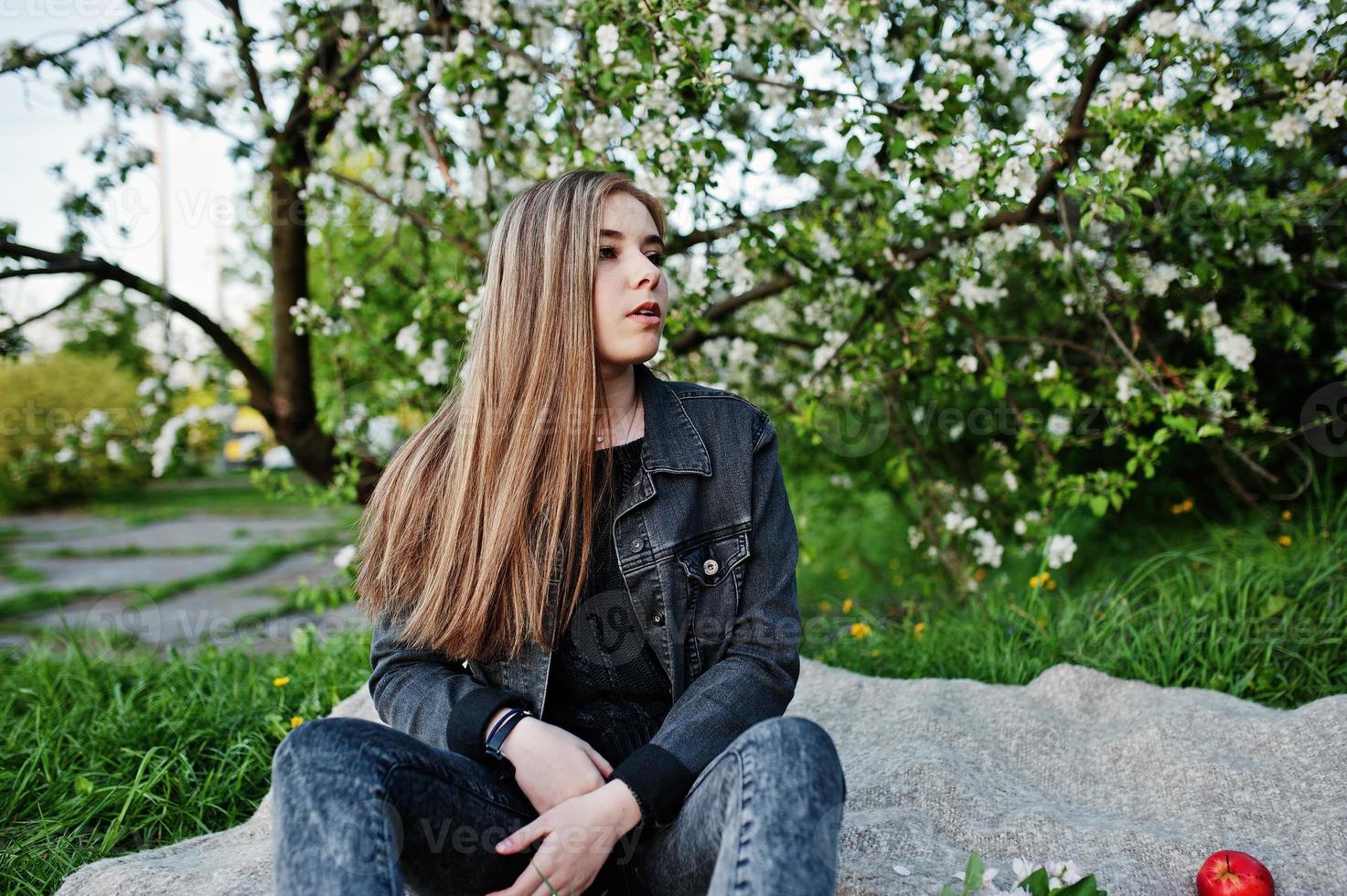 jovencita morena en jeans sentada en tela escocesa contra el árbol de flores de primavera. foto