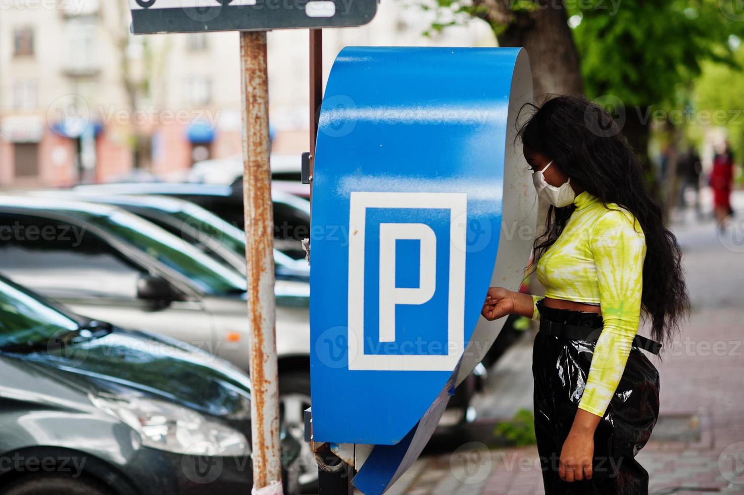 African American woman posing with facial mask to protect from infections from bacteria, viruses and epidemics, using parking pay station terminal. photo