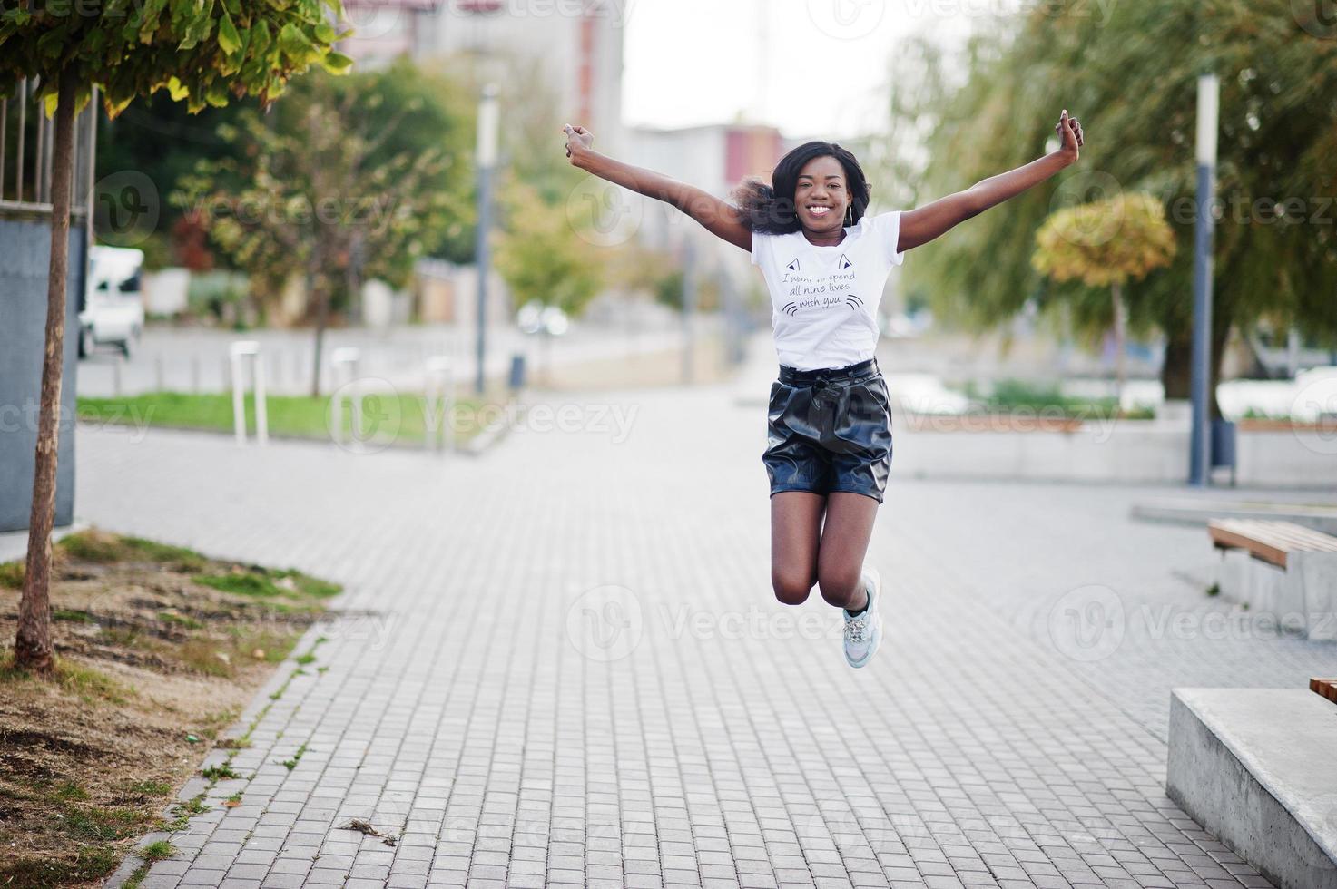 Modelo delgado afroamericano de piel oscura posado en pantalones cortos de cuero negro y camiseta blanca. ella saltando en el aire. foto