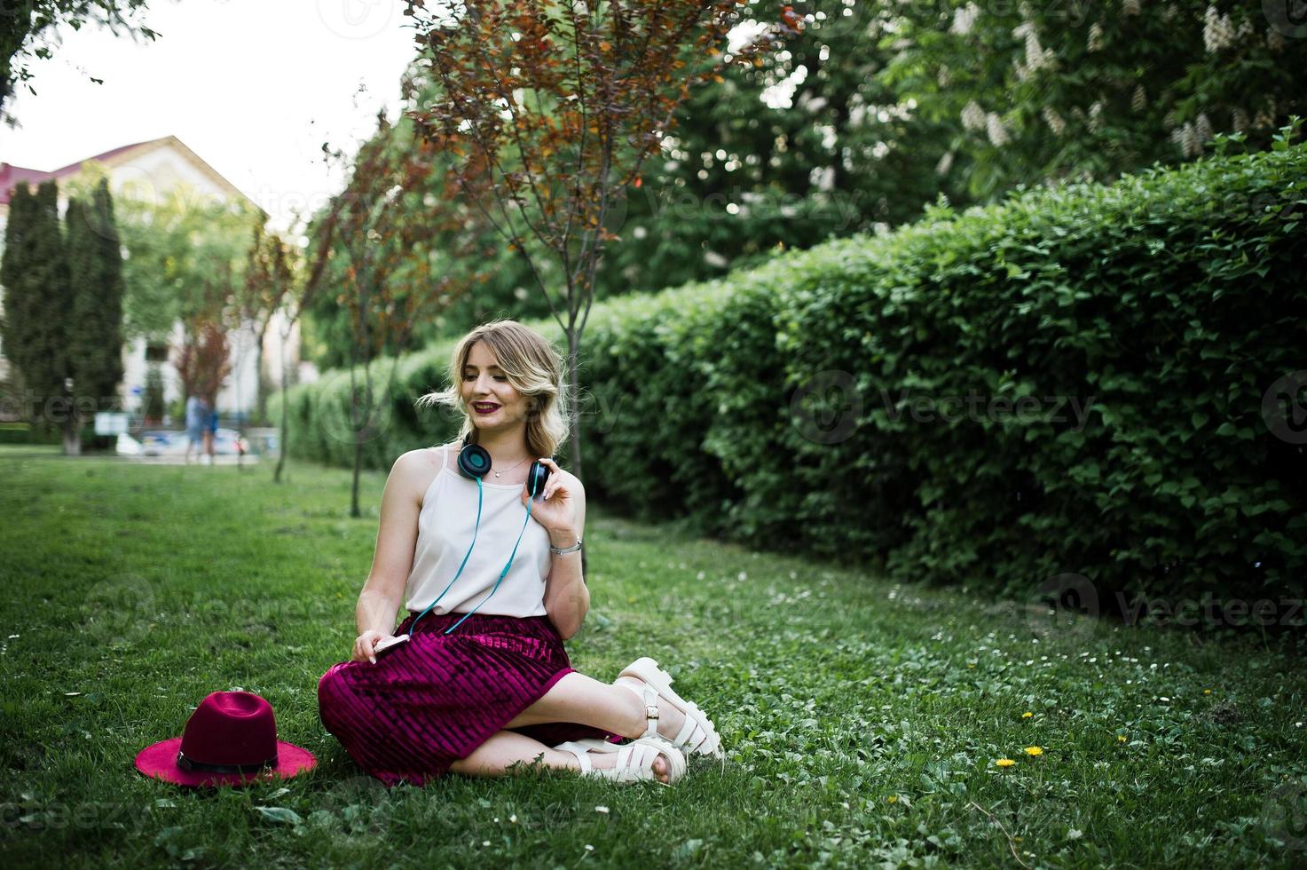 Fashionable and beautiful blonde model girl in stylish red velvet velour skirt, white blouse and hat, sitting on green grass at park with phone and earphones. photo