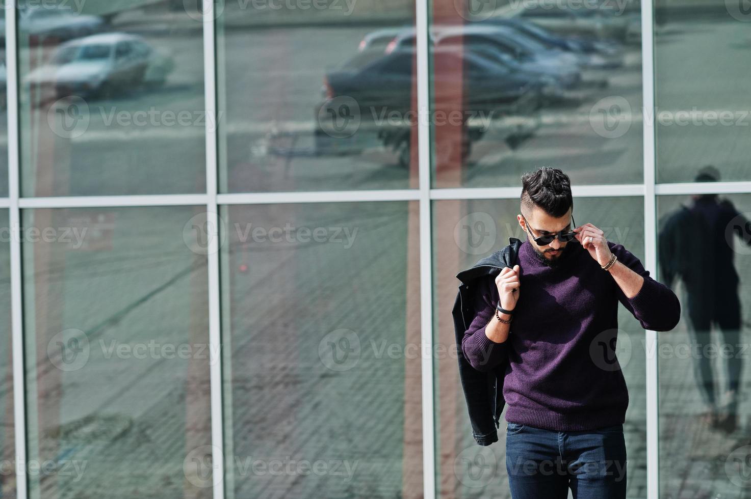 Fashion rich beard Arab man wear on turtle neck and sunglasses posed against large windows of modern building with jacket on hand. Stylish, succesful and fashionable arabian model guy. photo