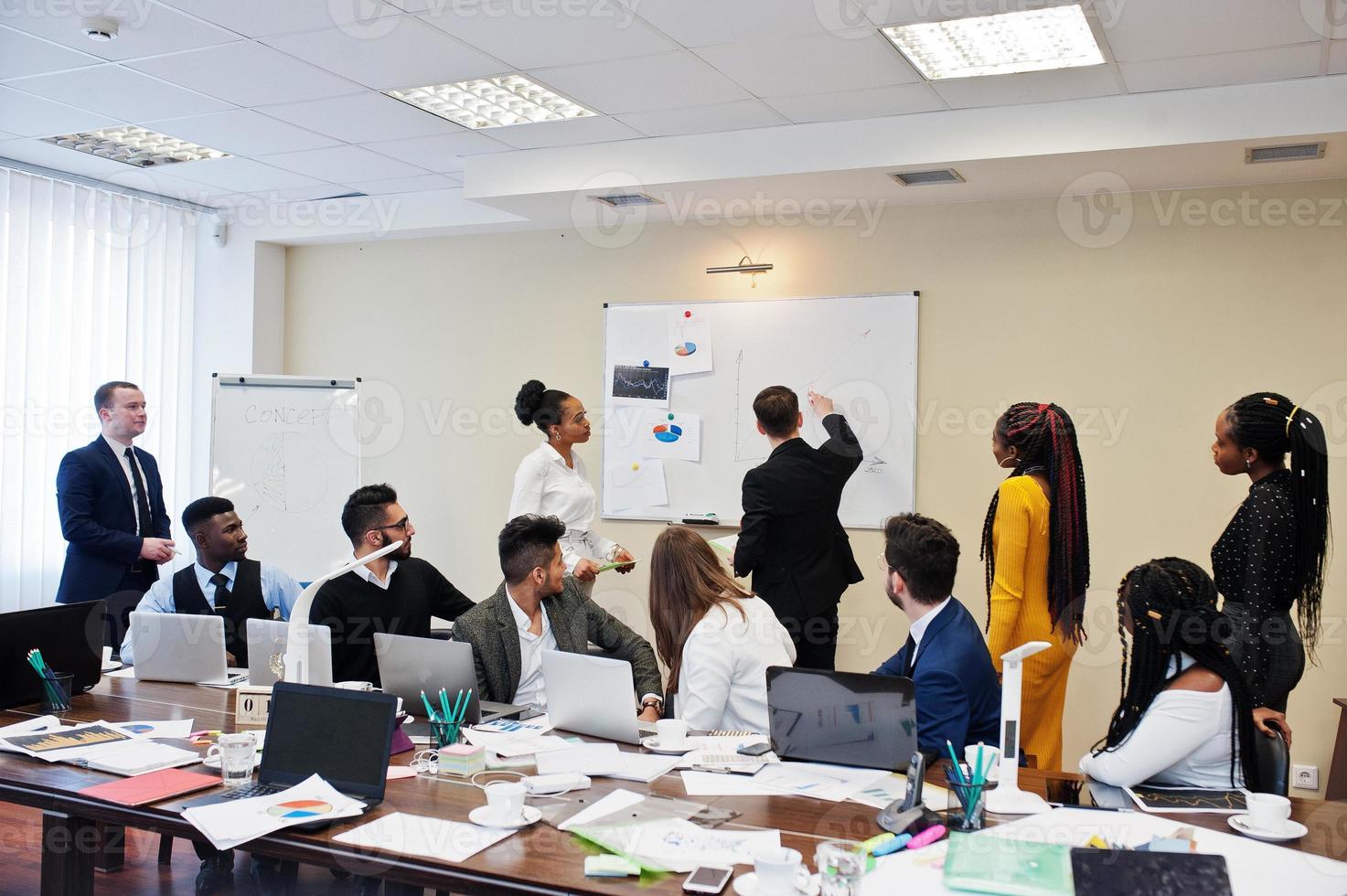 Mixed race business coach presenting report standing near whiteboard pointing on sales statistic shown on diagram and chart teach diverse company members gathered together in conference room. photo