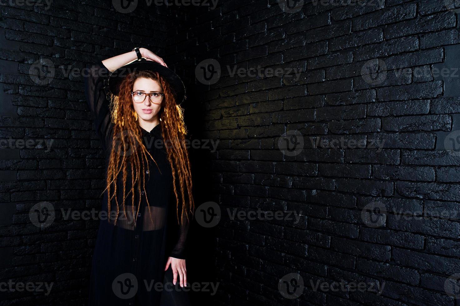 Studio shoot of girl in black with dreads, at glasses and hat on brick background. photo