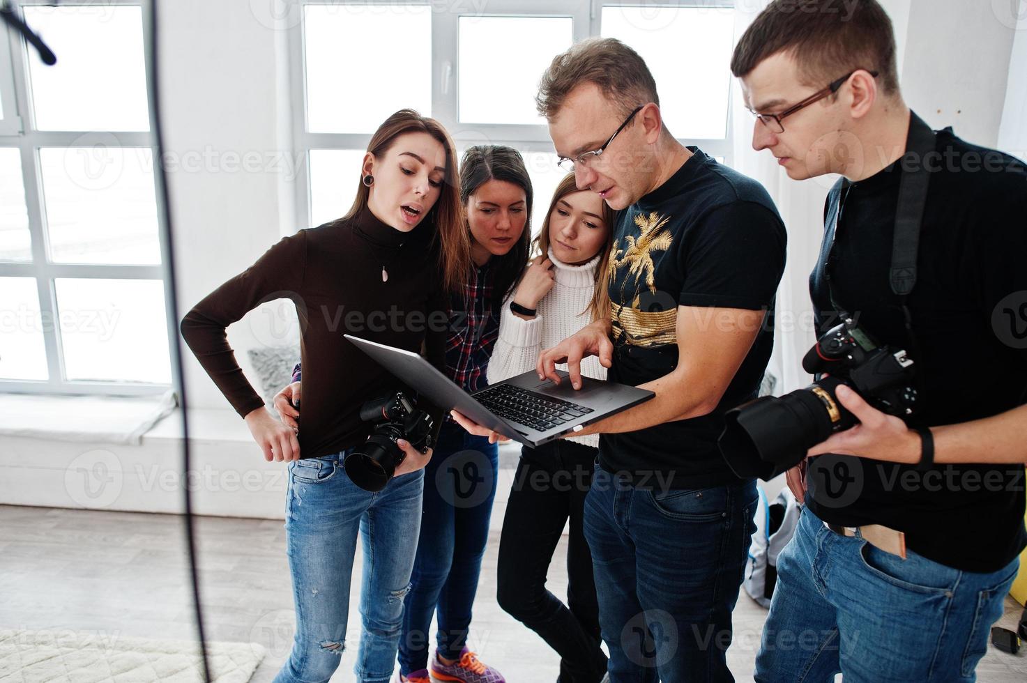 Photographer explaining about the shot to his team in the studio and looking on laptop. Talking to his assistants holding a camera during a photo shoot. Teamwork and brainstorm.
