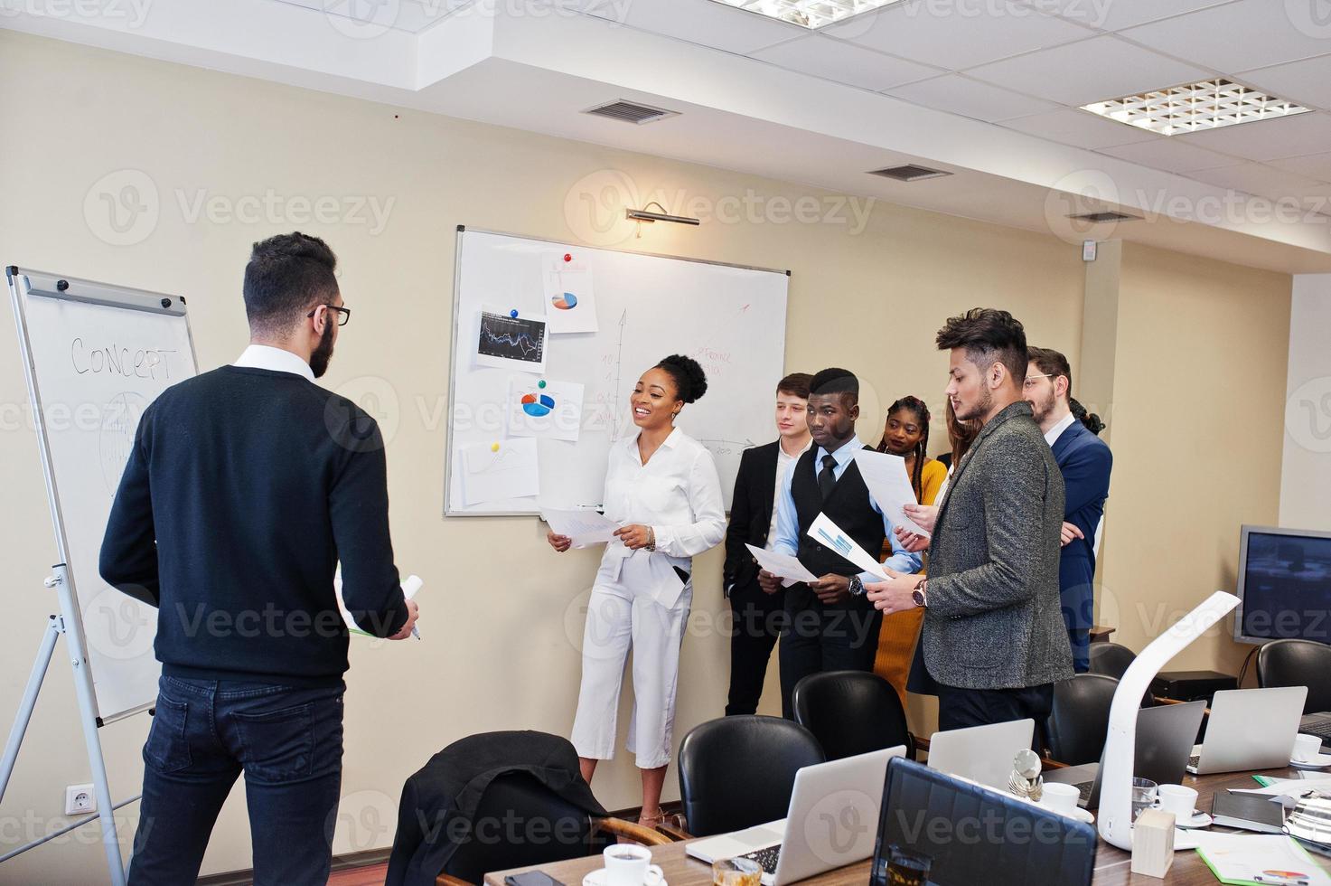 Mixed race business coach presenting report standing near whiteboard pointing on sales statistic shown on diagram and chart teach diverse company members gathered together in conference room. photo